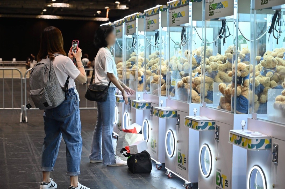 會展場館內一排八部白色品牌訂製夾公仔機,機內載滿藍衣公仔,兩名參加者站在機前